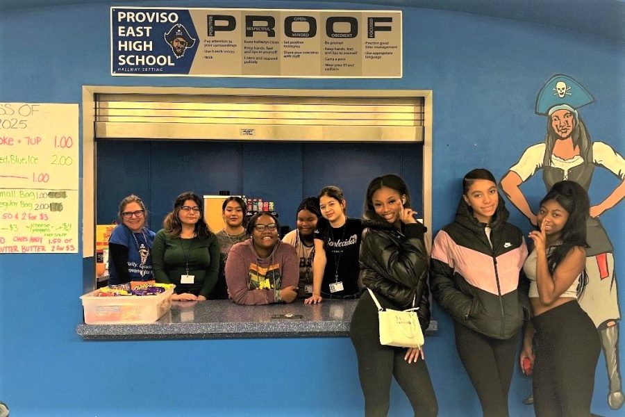 Three customers pose outside of the concession booth while the Class of 2025 members stand in the concession window. Inside the window, from left to right: Teacher/Sponsor Christine Corso, and students Yajaira Figueroa, Jocelyn Nambo, MyKal Westbrook, Deanna Adams, and Karen Lara.
